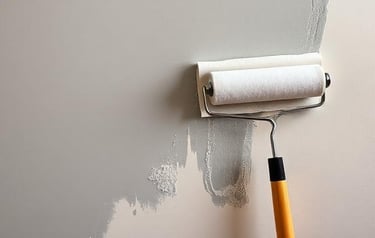 A white paint roller applying a fresh coat of light gray interior paint to a neutral colored wall.