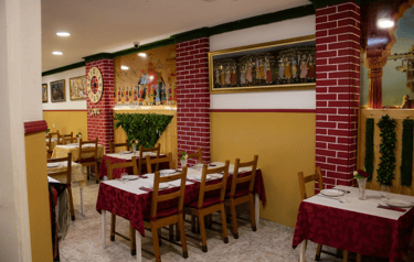 Indian restaurant interior with red patterned tablecloths, wooden chairs, and traditional murals on yellow walls.