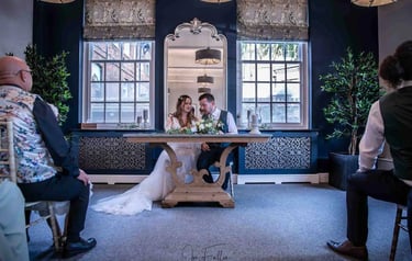 A bride and groom signing the marriage register during an indoor wedding ceremony with dark blue walls.