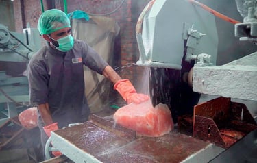 Worker cutting a large salt block with a machine in a factory, wearing safety gear and gloves.