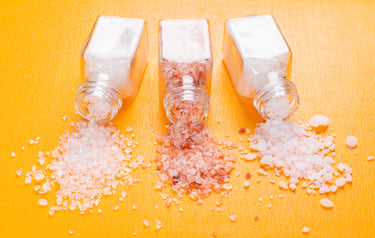 Three glass jars spilling different types of Himalayan salt granules on a bright orange surface