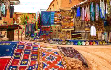 Handwoven Moroccan Berber rugs and colorful textiles displayed at an outdoor market in a clay village.