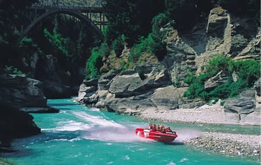 A red Shotover Jet boat speeding through the turquoise Shotover River canyon in Queenstown, New Zealand.
