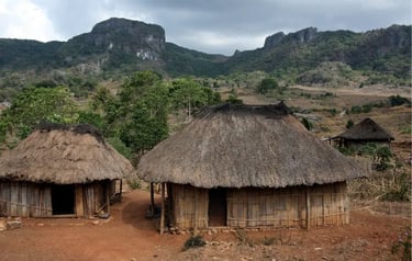 Traditional thatched roof huts in a rural mountain village landscape under a cloudy sky.