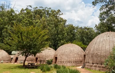 Traditional beehive-style thatched huts in a lush African village setting with green trees.