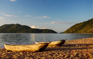 Two wooden dugout canoes rest on the sandy shores of Lake Malawi with green mountains in the distance.