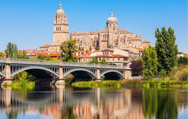 Salamanca Cathedral and the Roman Bridge reflecting in the Tormes River under a clear blue sky.