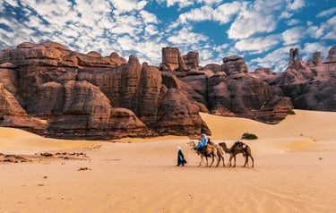 A camel caravan guided by people through the Sahara Desert sand dunes with rocky mountains.