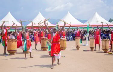 Burundi drummers in traditional red and white robes perform a rhythmic dance with wooden sticks.