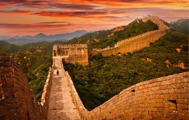 The Great Wall of China winding through green mountains under a vibrant orange sunset sky.