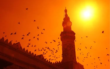Silhouette of a mosque minaret and a flock of birds flying during a bright orange sunset.