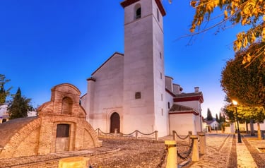 Historic white church and stone bell tower in a cobblestone plaza in Granada, Spain during twilight.