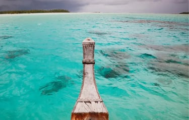 Traditional wooden boat bow sailing through clear turquoise tropical ocean waters under a cloudy sky.