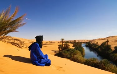 A man in traditional blue Tuareg clothing sitting on a Sahara Desert sand dune overlooking a lush river oasis.