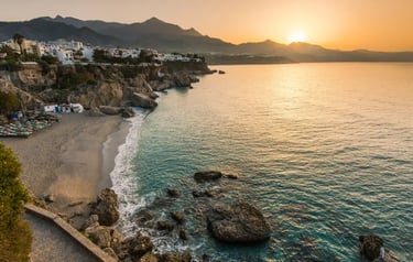 Sunset over the rocky coastline and sandy beach of Nerja, Spain, with mountains in the background.