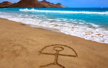 Indalo symbol drawn in the sand on a sunny beach in Almeria, Spain, with ocean waves and mountains.