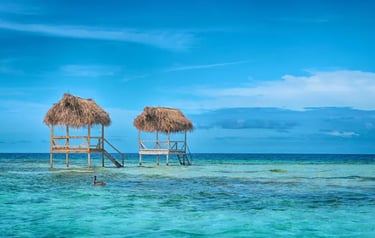 Two tropical thatch-roof huts standing in turquoise Caribbean waters under a bright blue sky.