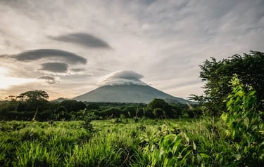 Scenic view of a tropical volcano peak topped with white lenticular clouds overlooking a green meadow.