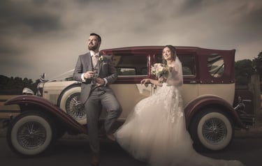 A bride and groom posing for wedding photos next to a vintage burgundy and cream classic car.