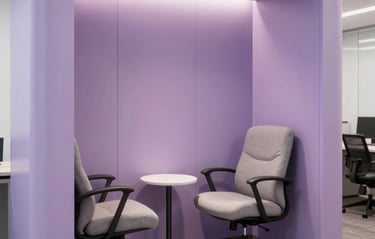 Photography of a custom corporate lactation booth interior in a modern workplace in Hispanic / Corporate Latin America & Spain. Features pastel lilac wall paneling, ergonomic armchair, soft accent illumination, side table, private ambient.