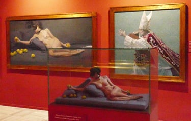 Contemporary art exhibition featuring a nude sculpture in a glass case beside classical oil paintings.