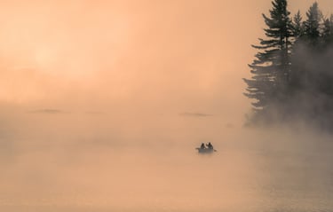 Two people rowing a canoe on a calm, misty lake at sunrise surrounded by pine trees.