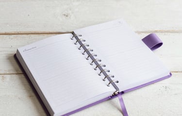 Flat lay of a beautifully crafted personalized creative planner and journal with lilac ribbons, lying on a clean whitewashed wooden surface in a Portuguese home design studio.