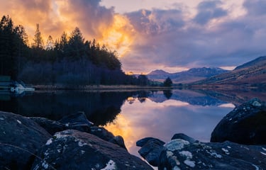 Scenic sunset over a calm lake with mountain reflections and rocky shoreline in the wilderness.