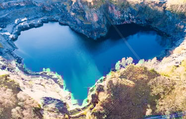 Aerial view of a heart-shaped blue lake in a rocky quarry surrounded by autumn trees.