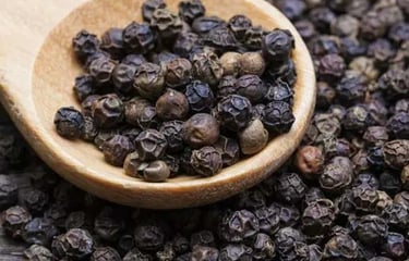 Organic whole black peppercorns in a wooden spoon over a pile of dry peppercorns.