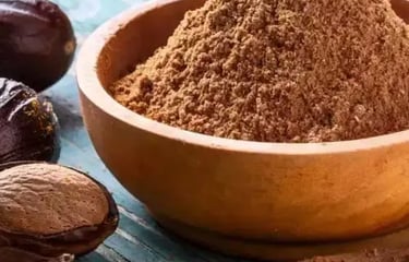 A small wooden bowl filled with ground nutmeg powder next to whole nutmeg seeds on a rustic blue table.