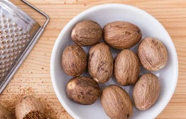 Whole nutmeg seeds in a white bowl next to a metal grater and freshly ground nutmeg on a wooden surface.