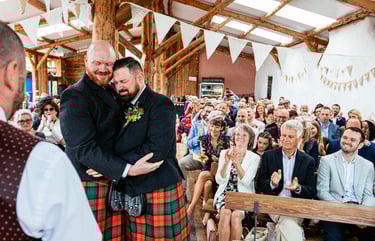 Grooms embrace during wedding ceremony in rustic barn at South Wales venue Ceridwen, with guests clapping and cheering