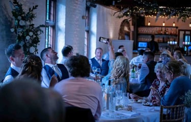 Wedding guests smiling and taking photos with smartphones at a rustic indoor reception dinner.
