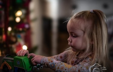 Toddler girl playing with a green toy truck near a blurred Christmas tree with lights. Athens, Oh