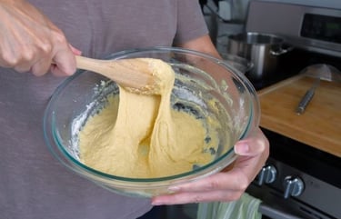 A person using a wooden spoon to mix thick yellow bread dough in a glass bowl.
