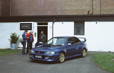 a classic Subaru Impreza parked in front a building with three guys looking at it
