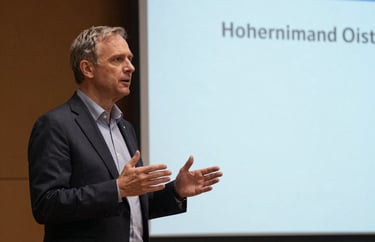 A speaker on stage at an educational conference, gesturing towards a presentation screen. North American / International academic setting.