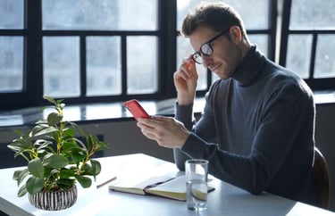 A man in glasses looks at his red smartphone at a bright desk with a potted green plant.