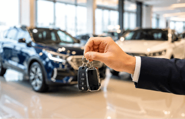 Mão segurando chave de carro em concessionária com veículos desfocados ao fundo.