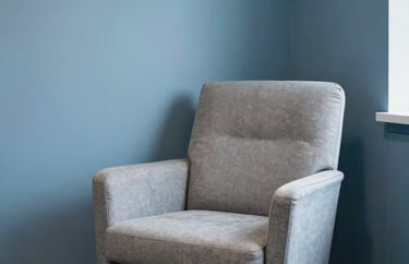 A comfortable, modern armchair in a light-filled corner of an office with soft steel blue walls, representing a safe space for individual therapy.