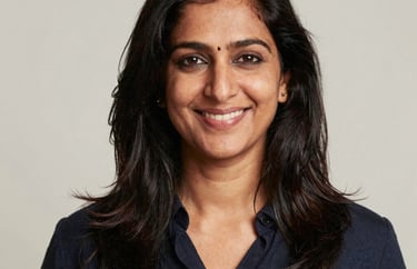 A professional studio portrait of an approachable South Asian woman smiling, set against a neutral off-white background. Professional lighting highlighting a trustworthy and warm persona.