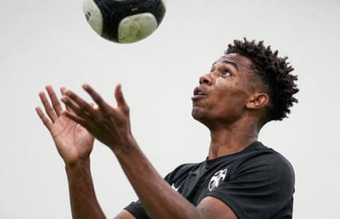 Action shot of a young male player of African descent, aged 15, performing a header. Sharp focus, clean white background of a professional facility wall. Intense commitment in his eyes.