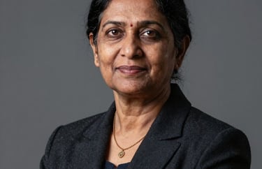 Professional studio portrait of a senior South Asian female executive with a friendly yet firm look, dark formal blazer, grey background.