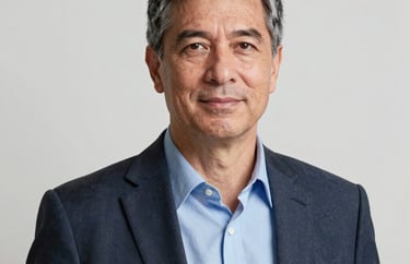 A professional headshot of a male senior coach in business casual attire, standing in front of a clean white wall with soft, flattering lighting.