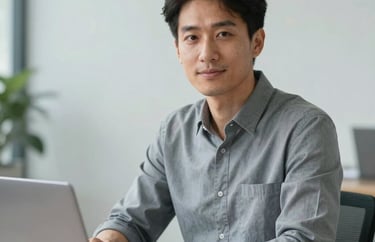 A professional headshot of a person working at a desk with a laptop, looking approachable and confident. The background is a clean North American office with light grey tones.