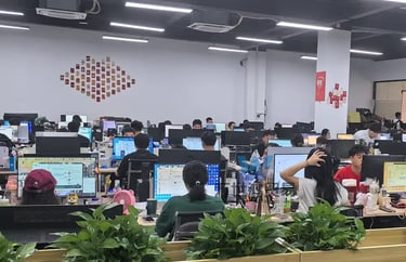 Employees working at computer desks in a modern open-plan office with green plants.