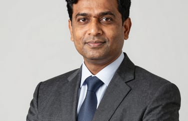 A professional studio portrait of a South Asian male educator in formal attire. Sharp lighting, clean professional aesthetic against a light background.