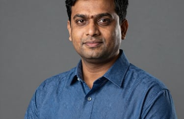 A professional studio portrait of a South Asian male teacher in a blue formal shirt. He looks approachable and authoritative. Neutral gray background.