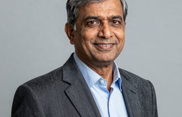 A professional studio portrait of a senior South Asian male educator wearing a formal blazer, smiling confidently. The lighting is professional and the background is a solid light gray.